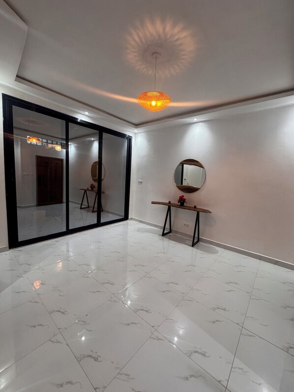 A spacious room with a large sliding glass door, a round mirror on a wooden stand, and a modern ceiling light fixture.