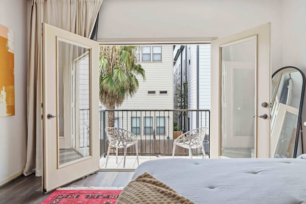 Une chambre avec un grand lit, un balcon avec des chaises et une vue sur les immeubles.