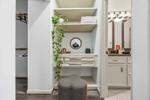 Une salle de bain avec un meuble-lavabo blanc, un miroir, un porte-serviettes, une plante et un petit tabouret.