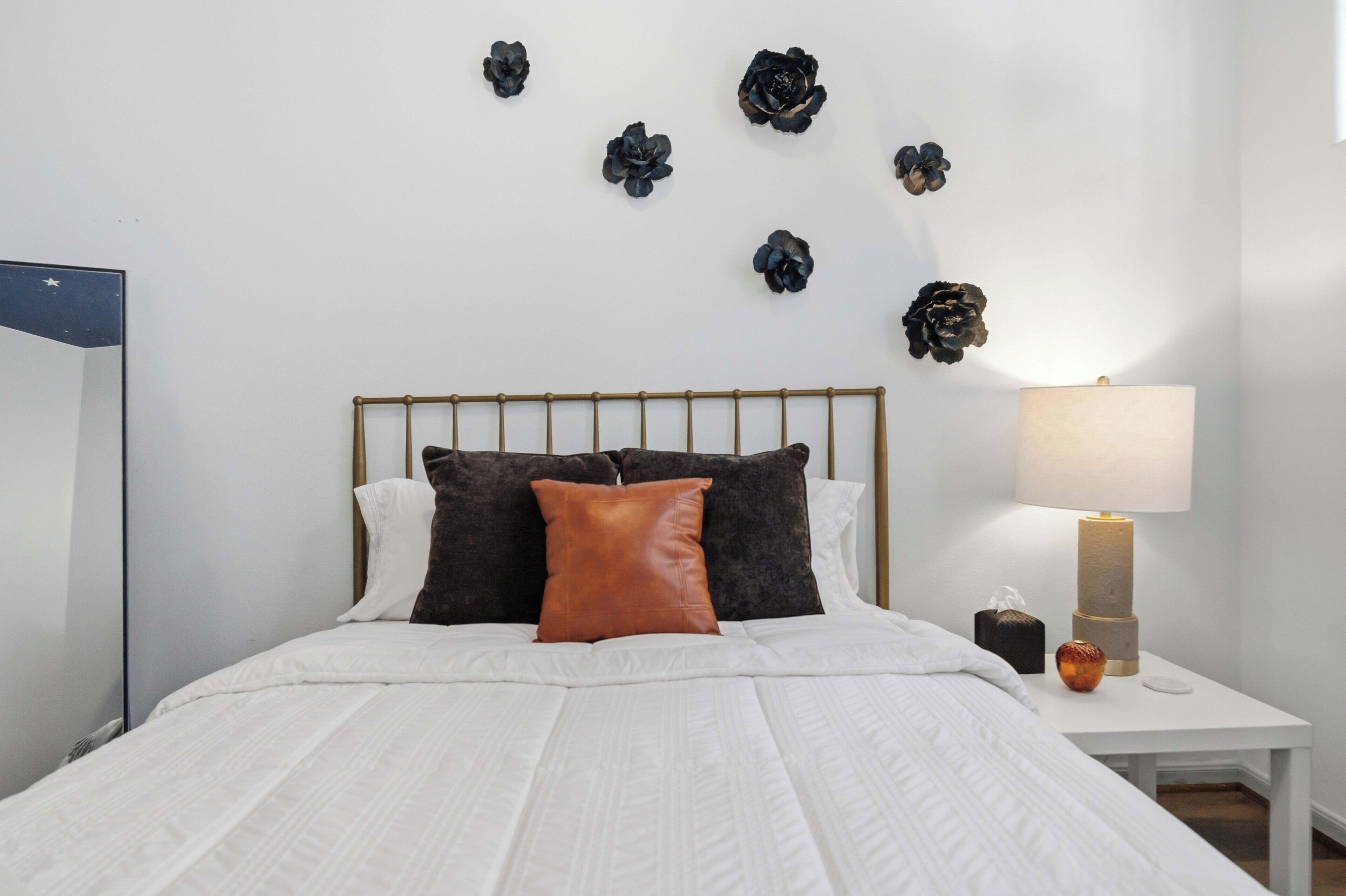 Ein ordentlich bezogenes Bett mit weißer Decke und braunem Kopfkissen, ein Nachttisch mit Lampe sowie Wanddeko.