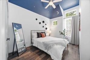 Une chambre à coucher avec un lit, un ventilateur de plafond, une fresque murale représentant un ciel étoilé, une fenêtre et un miroir.