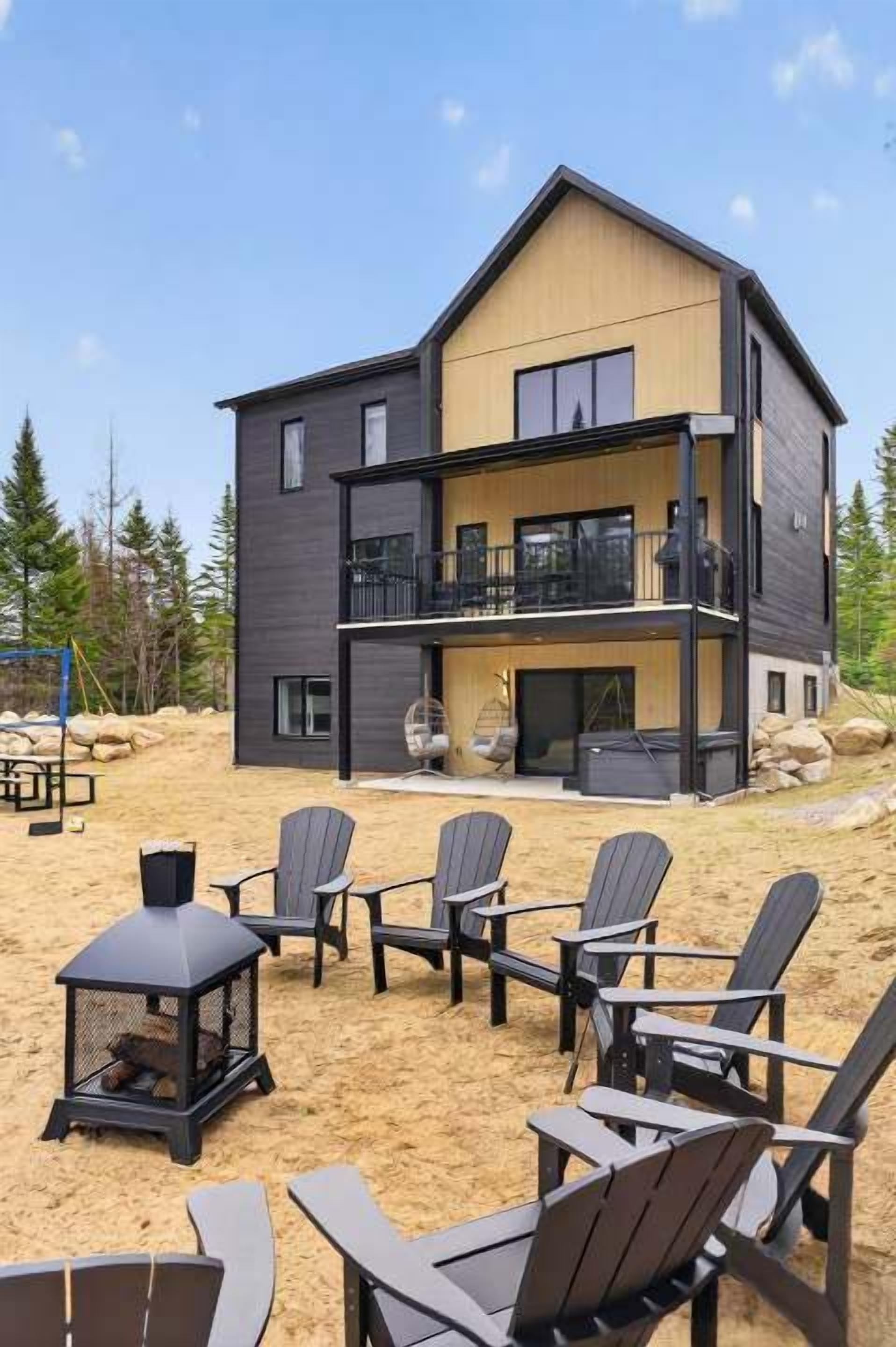 A modern two-story house with a balcony, outdoor seating, and a wood-burning stove.