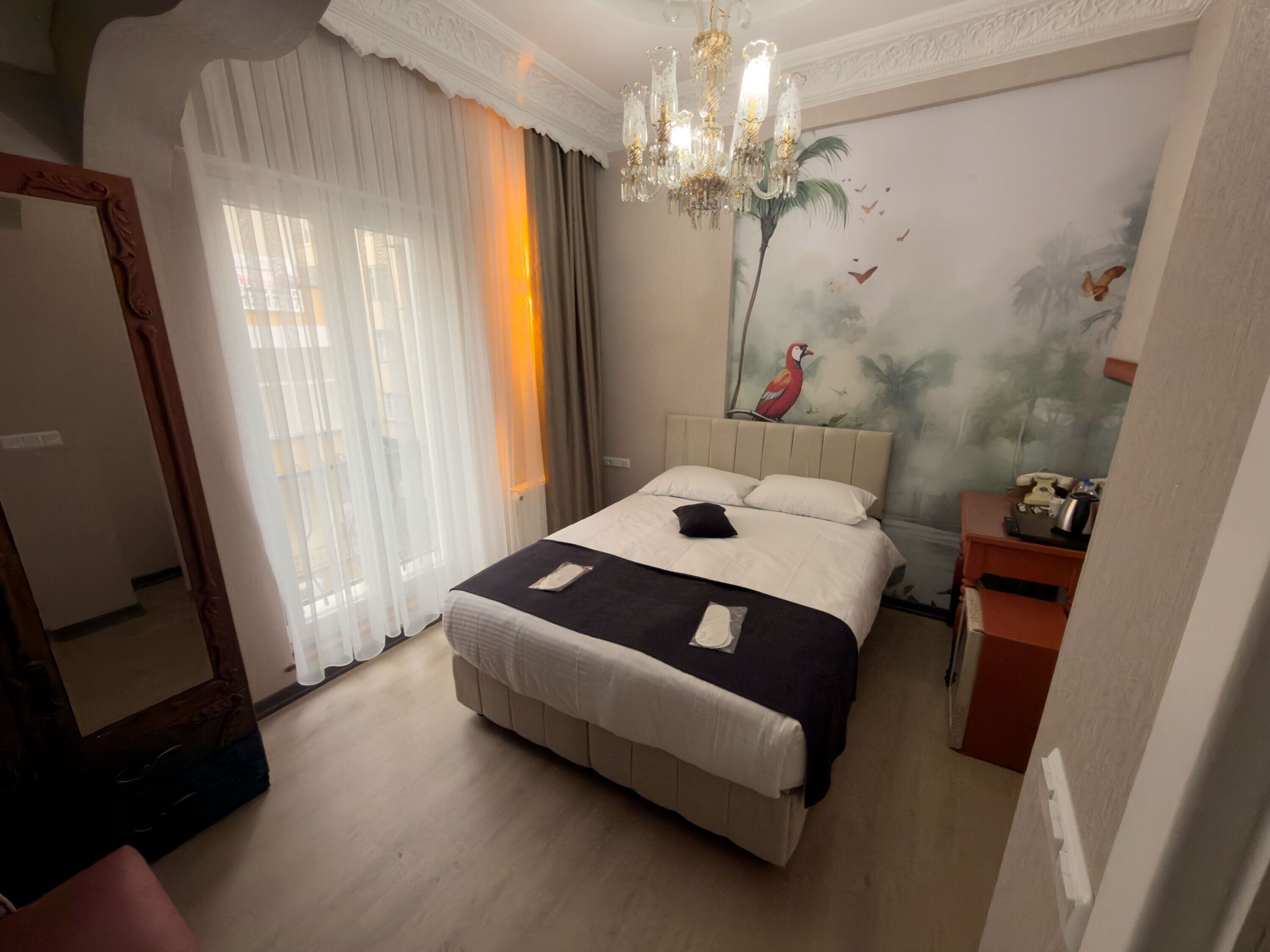 A hotel room with a large bed, a chandelier, a painting of a bird on the wall, and a telephone on a side table.