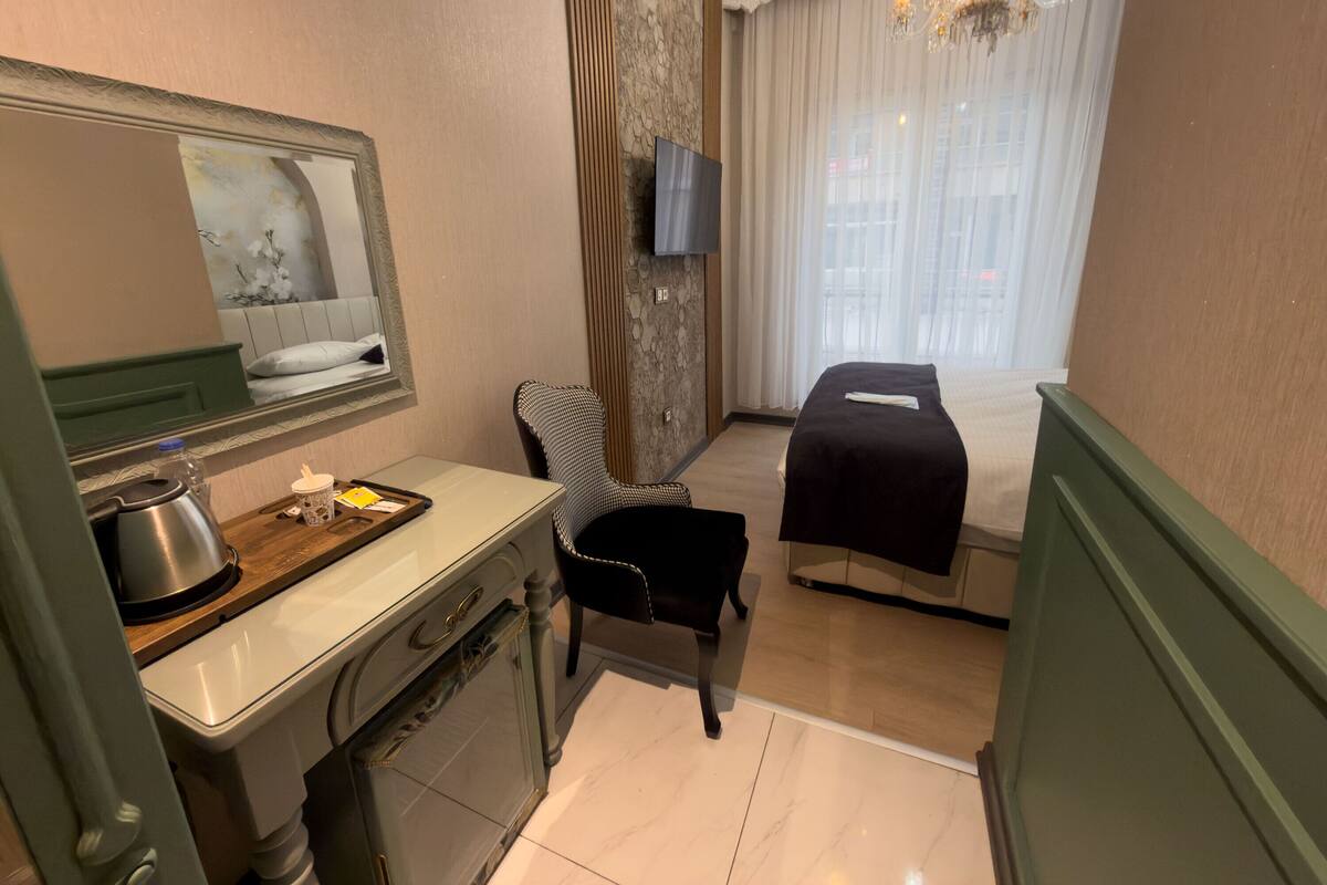 A hotel room with a bed, a chair, a desk with a kettle, a mirror, and a TV.