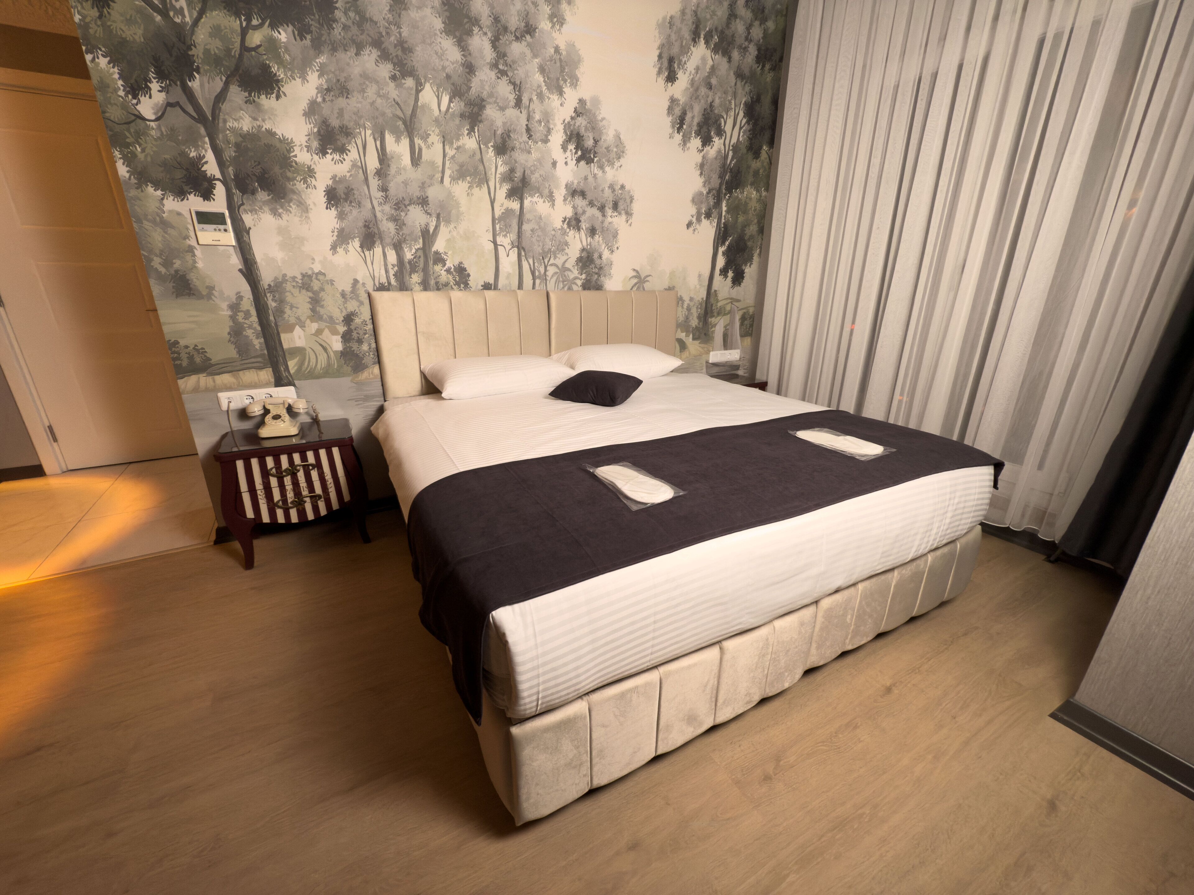A neatly made bed with a headboard, a bedside table with a telephone, and a wall mural of trees.