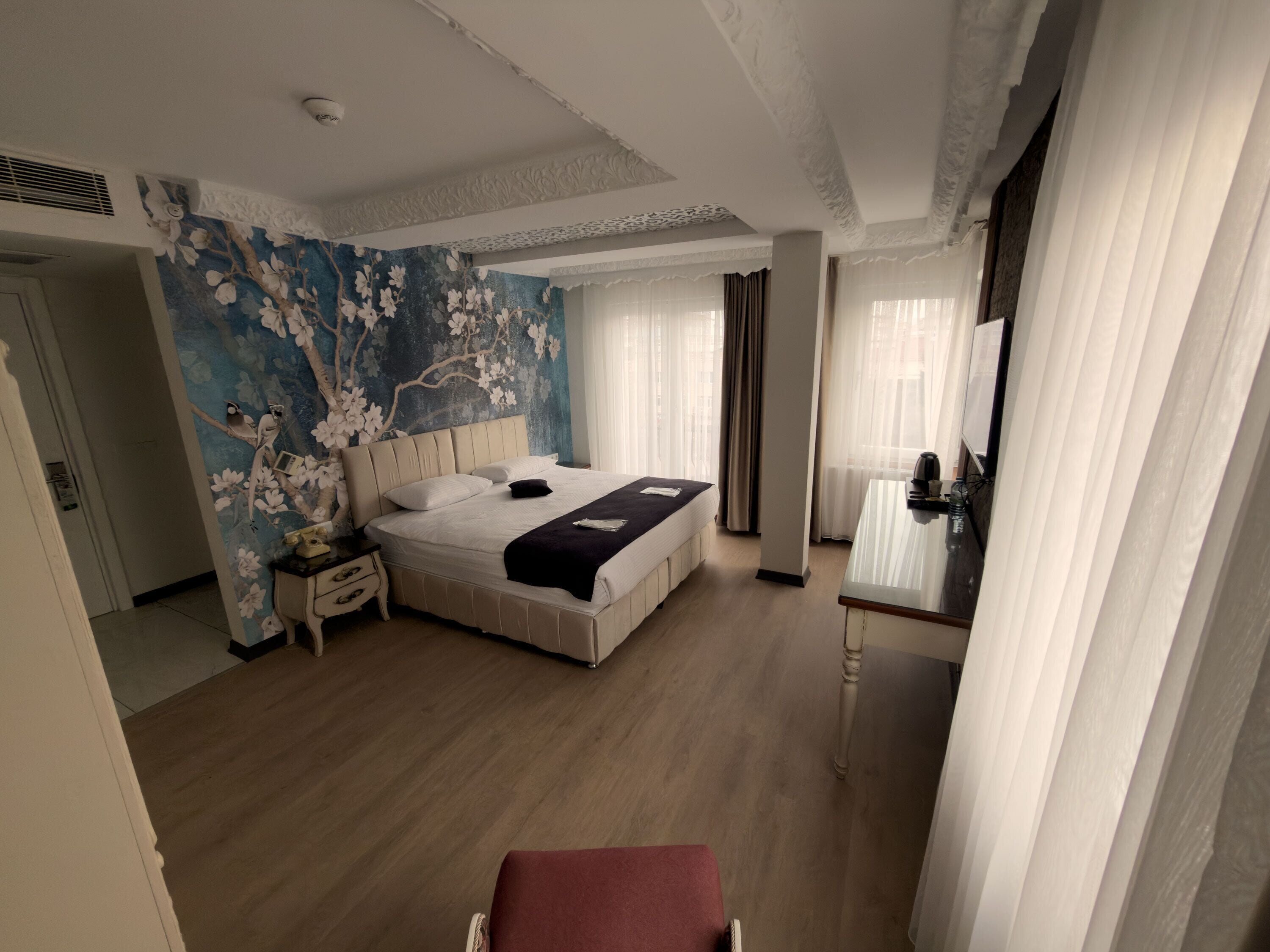 A hotel room with a large bed, a nightstand, a desk with a chair, a mirror, and a wall with floral wallpaper.