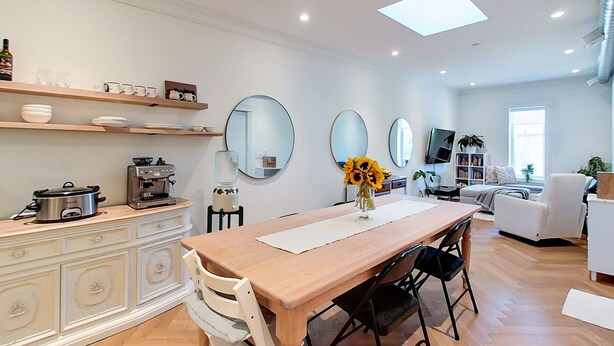 A modern kitchen with a wooden table, white cabinets, and a coffee machine. There are two mirrors on the wall, a TV, and a bookshelf with books and plants. - Chic Retreat Downtown Meaford 2 min to Georgian bay and ~20 min to ski hills (Meaford)