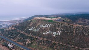 Eine Vogelperspektive zeigt eine hügelige Landschaft mit arabischen Schriftzügen, einer Straße und einer entfernten Küstenlinie.