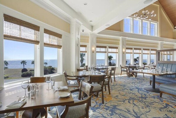 A dining area with tables set for service, large windows with views of the ocean, and a high ceiling with a chandelier.