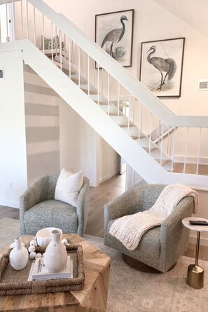 Una escalera con barandas blancas, dos sillones grises con cojines blancos, una mesa de centro de madera con adornos y cuadros en la pared con motivos de pájaros.