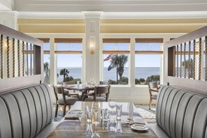 Comedor con vista al océano, con una mesa dispuesta con vasos y platos, y asientos acolchados.
