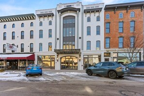 A multi-story building with a central entrance, storefronts on the ground floor, and a parking lot with cars. - Local Discounts-Charming View of Downtown Newark (Newark)