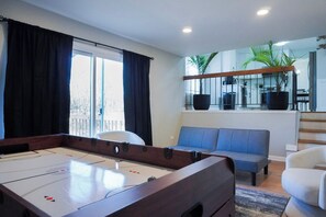 Een kamer met een tafelvoetbaltafel, een blauwe bank en een trap naar een hoger gelegen niveau met een balkon.