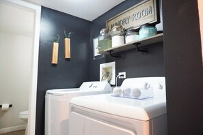 A laundry room with a washing machine, dryer, shelves with storage containers, and a sign indicating the room's purpose.