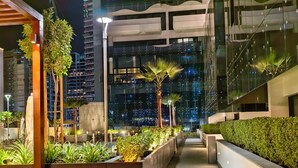 Un espacio urbano exterior iluminado de noche, con edificios modernos, jardineras con follaje y un camino bien iluminado.