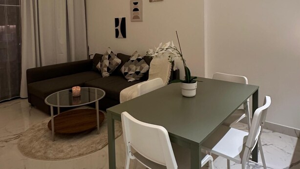Una sala de estar moderna con una mesa de comedor, sillas blancas, un sofá con cojines estampados, una mesa de centro de vidrio y un reloj en la pared.