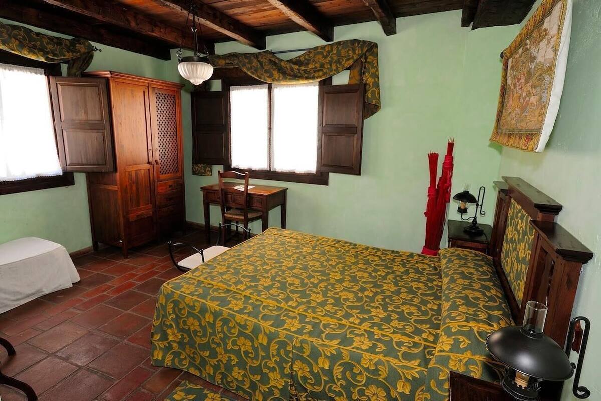Een kamer met een bed, een bureau en een stoel. Er zijn twee ramen met gordijnen, een houten garderobe en een wanddecoratie.