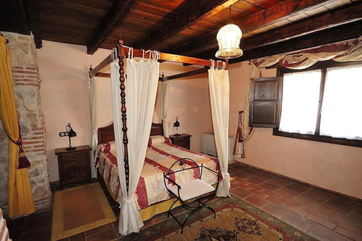 Een hemelbed met witte gordijnen, een houten plafond en een stenen muur.