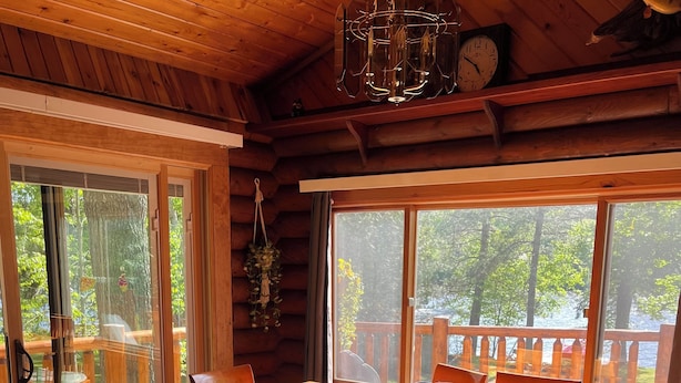 A wooden cabin with a dining area featuring a large table and chairs, a chandelier, and a view of a deck and trees through large windows. - Wisconsin River Getaway. Large yard and shoreline. Pet friendly. (Merrill)