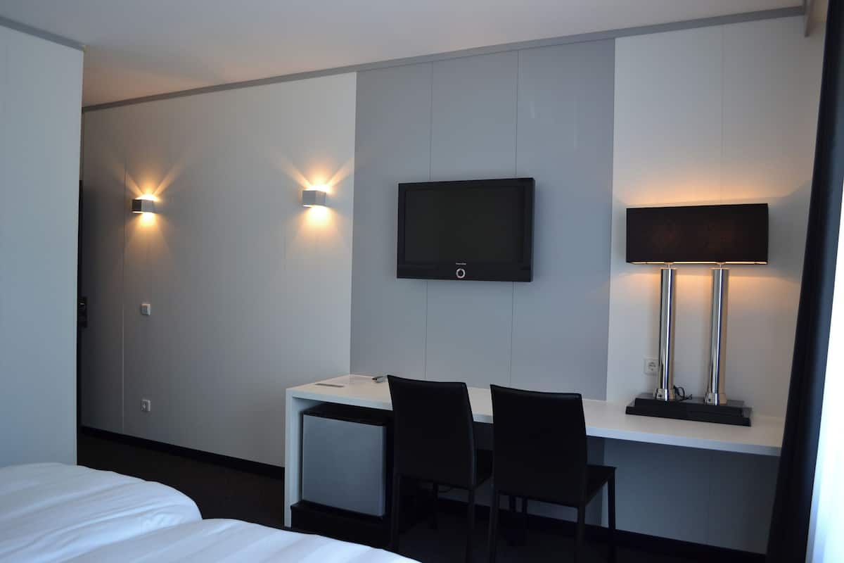 A hotel room with a bed, a desk, two chairs, a TV mounted on the wall, and two wall-mounted lamps.