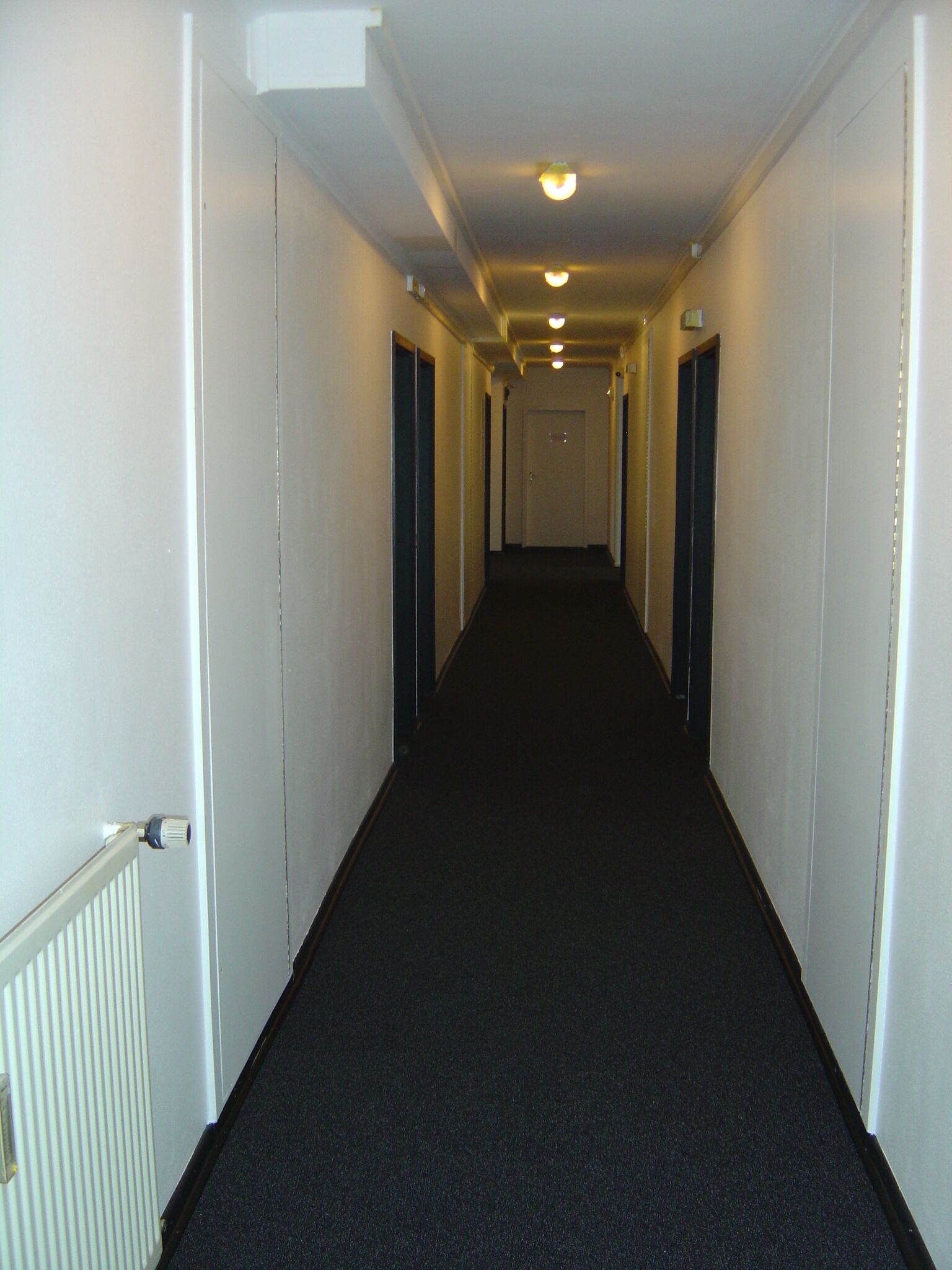 A long, narrow hallway with white walls and a dark carpet.