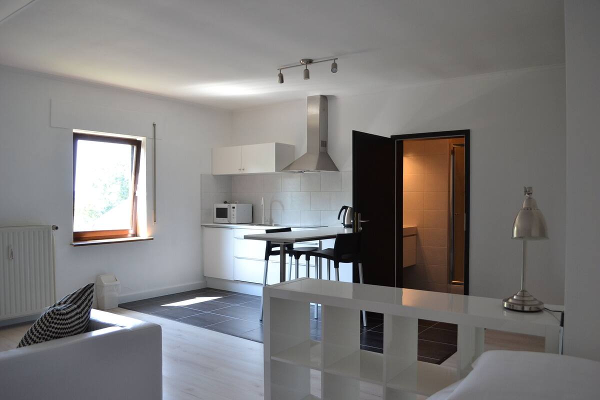 A modern kitchen with white cabinets, a microwave, and a stove. There is a dining area with a table and chairs, and a window providing natural light.