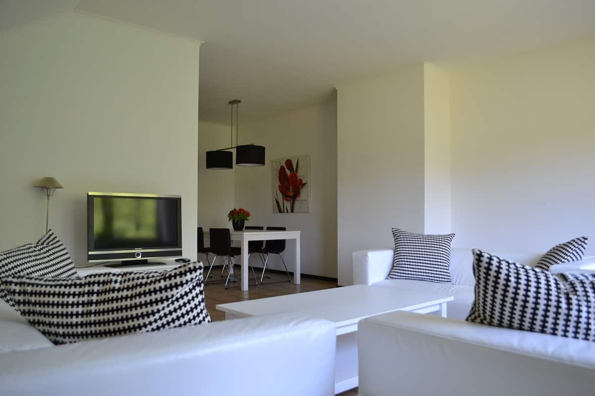 A modern living room with white furniture, a flat-screen TV, and a dining area with a white table and chairs.