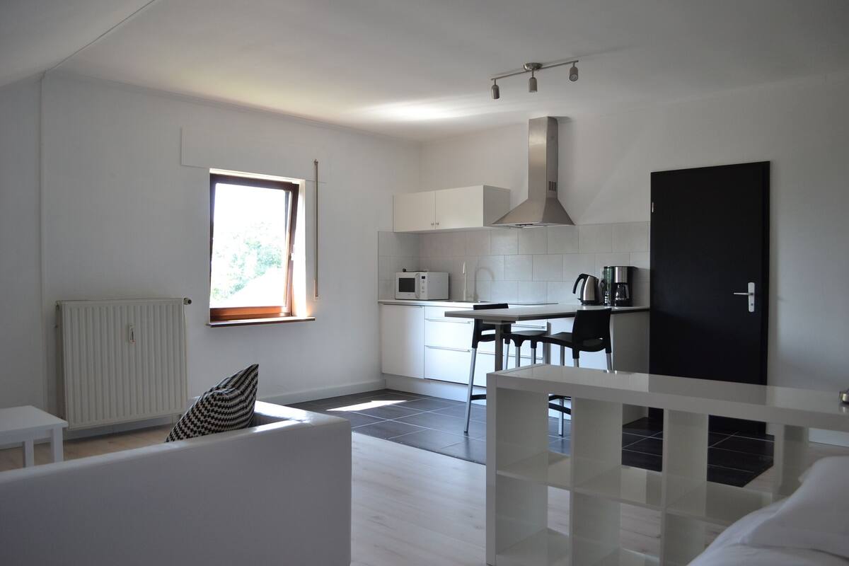 A modern kitchen with white cabinets, a microwave, and a stove. There is a dining area with a table and chairs. A black door is visible on the right side.