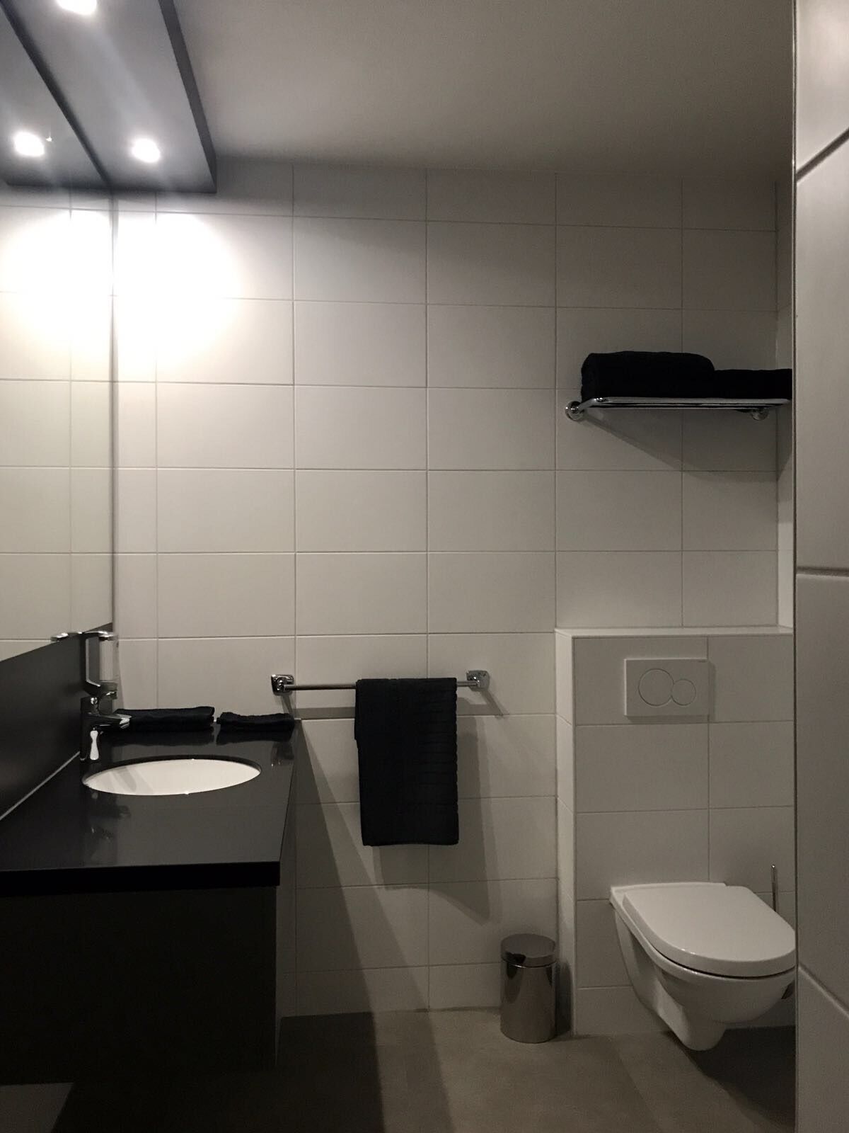 A modern bathroom with a black sink, white tiles, a towel rack, a toilet, and a trash can.