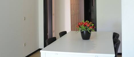 A modern dining area with a white table, black chairs, and a black pendant light. A vase with red flowers is on the table.