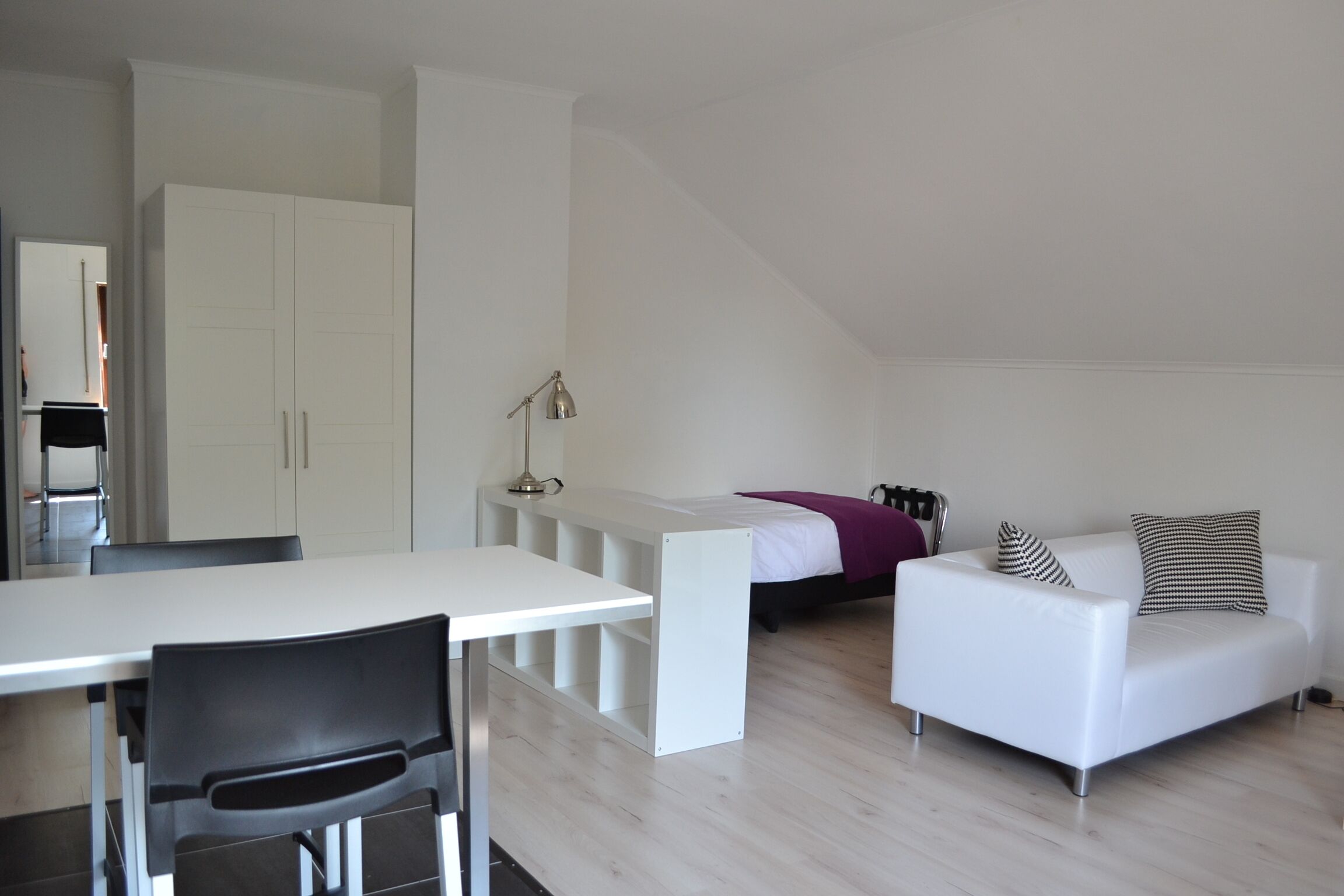 A modern, minimalistic room with a white desk, black chair, white sofa, and a bed with a purple blanket.