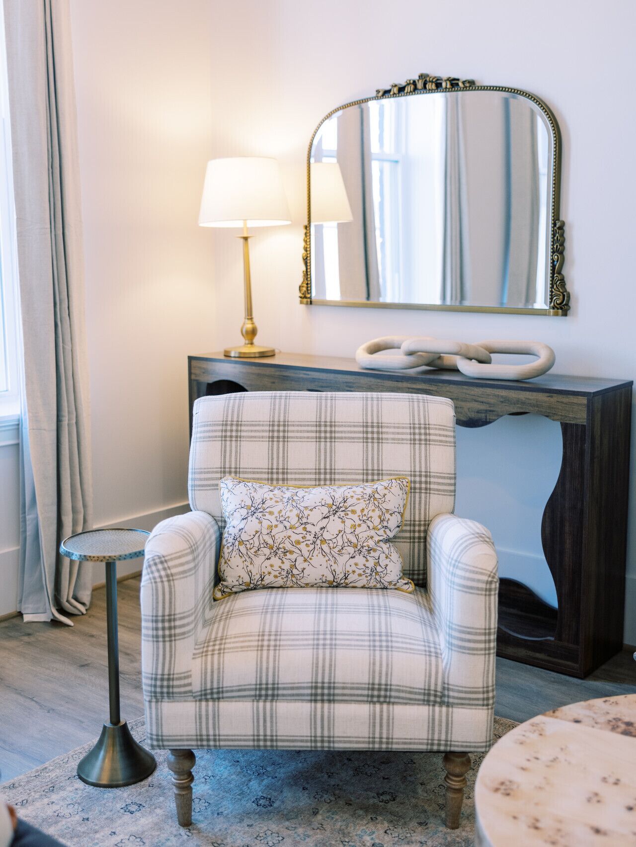 Un fauteuil à carreaux avec un coussin à motifs floraux, une table ronde, un lampadaire, un miroir et une console en bois foncé.