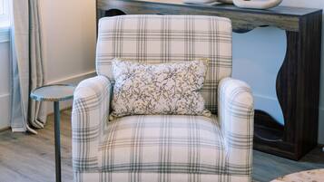 Un fauteuil à carreaux avec un coussin à motifs floraux, une table ronde, un lampadaire, un miroir et une console en bois foncé.