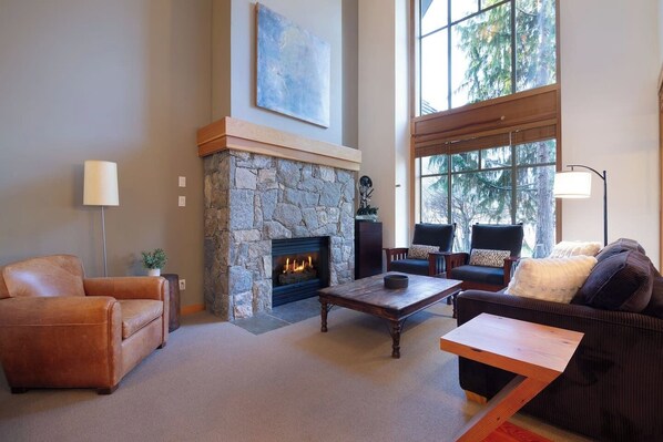 A living room with a stone fireplace, leather sofas, and a large window offering a view of trees. - Charming 4BR/5BA on Golf Course w/ Private Garage (Whistler)