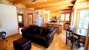 A cozy living room with a leather sofa, wooden flooring, and a kitchen area with wooden cabinets and a dining table. - Unforgettable Moments at Lake Pohénégamook (Pohénégamook)