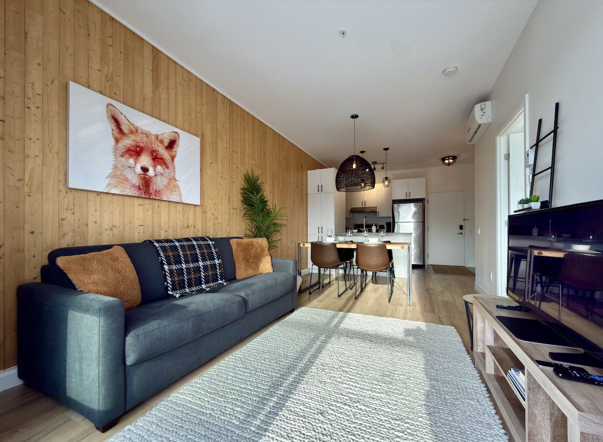 Una sala de estar moderna con un sofá gris, una pared de madera y una cocina al fondo.