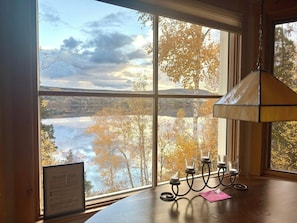 A room with a large window overlooking a body of water and trees, a table with a candle holder, and a framed notice on the window sill. - Serenity Overlooking Lake Pohénégamook (Pohénégamook)