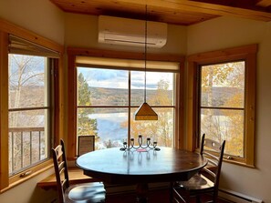 Una mesa redonda para comer con un candelabro, rodeada de sillas, en una habitación con ventanas amplias que dan a un lago y árboles.