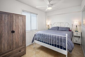 Ein Schlafzimmer mit einem Bett, einem Kleiderschrank, einem Deckenventilator, zwei Nachttischen mit Lampen und einem Fenster mit Jalousien.