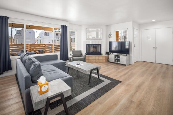 Ein modernes Wohnzimmer mit einem grauen Sofa, einem Couchtisch und einem Fernseher. Ein Kamin, ein Fenster mit Vorhängen und ein Blick auf Gebäude sind zu sehen.