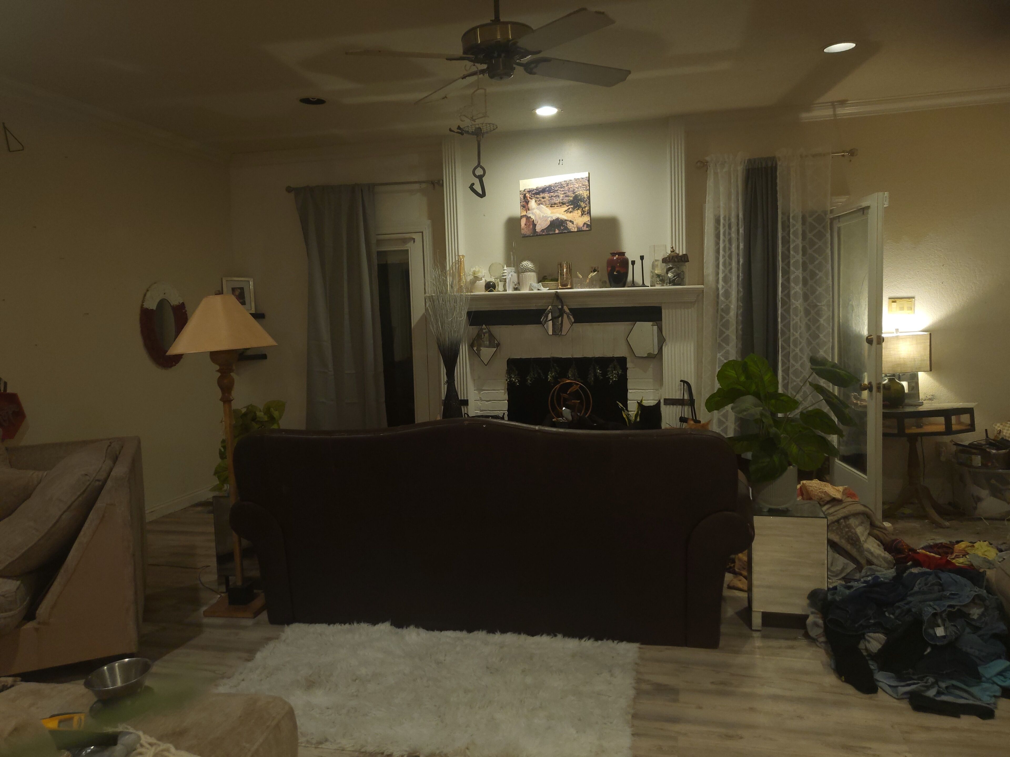 Uma sala de estar com um sofá escuro, um tapete branco e fofo, um ventilador de teto, uma lareira com ajeira e uma mesa desorganizada com diversos objetos.