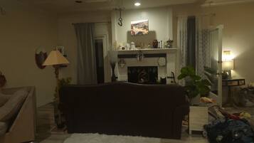 Uma sala de estar com um sofá escuro, um tapete branco e fofo, um ventilador de teto, uma lareira com ajeira e uma mesa desorganizada com diversos objetos.