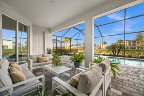 A spacious outdoor patio with a pool, white furniture, and large windows offering views of palm trees and buildings. - Charming lakeside home with pool, spa  and modern coastal design. (Ave Maria)