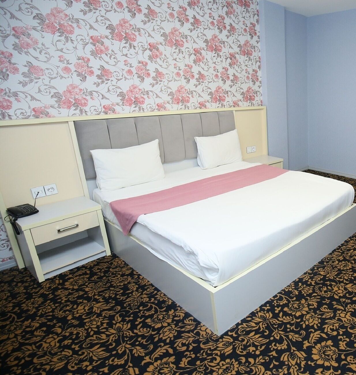 Hotellihuone, jossa on sänky, yöpöytä, pistorasia ja kuviollinen matto.