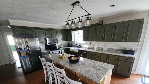A modern kitchen with granite countertops, green cabinets, a stainless steel refrigerator, and a bar area with white chairs.