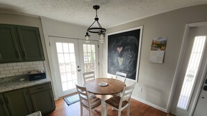 A kitchen with a wooden dining table, white chairs, a black and white abstract painting, and a calendar on the wall.