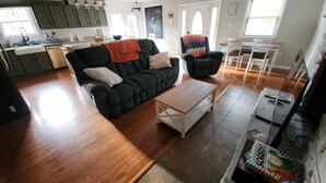 A spacious living area with a dark grey sofa, a wooden coffee table, and a black armchair with a colorful blanket. A kitchen with green cabinets and a dining area with a white table and chairs are visible.