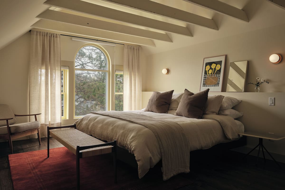 Un dormitorio con una cama grande, un banco, una silla, una ventana con cortinas y un cuadro en la pared.