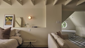 Una habitación acogedora con una cama, un sofá, una mesita y una estantería con un jarrón y un cuadro enmarcado.