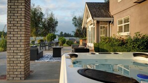Un jacuzzi en un patio trasero con un pilar de ladrillo, mobiliario exterior y una casa al fondo.